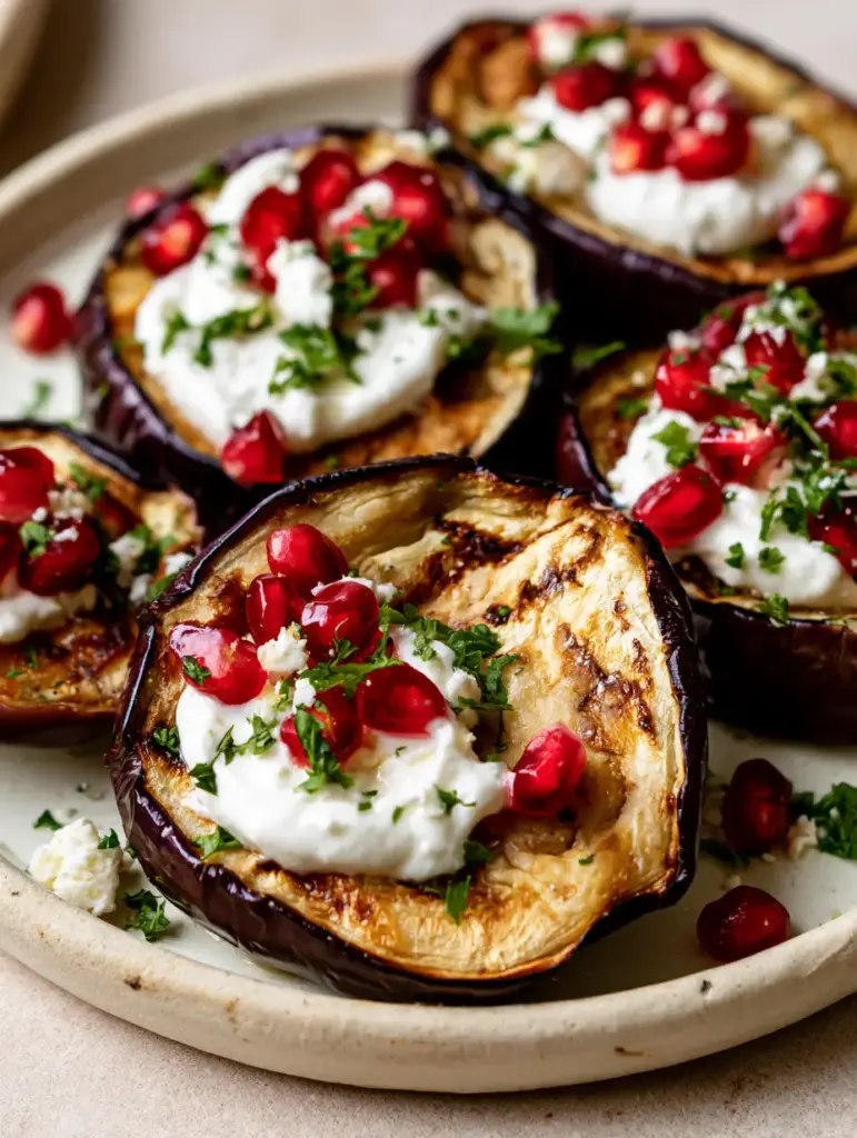 Greek Roasted Eggplant with Creamy Yogurt and Pomegranate 1 A rustic off-white ceramic plate filled with Greek Roasted Eggplant rounds garnished with parsley and pomegranate.
